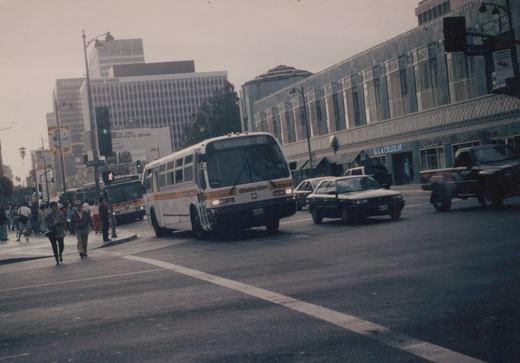 A white and orange Metro Bus is turning at a city intersection with pedestrians crossing and cars nearby, under a cloudy sky.