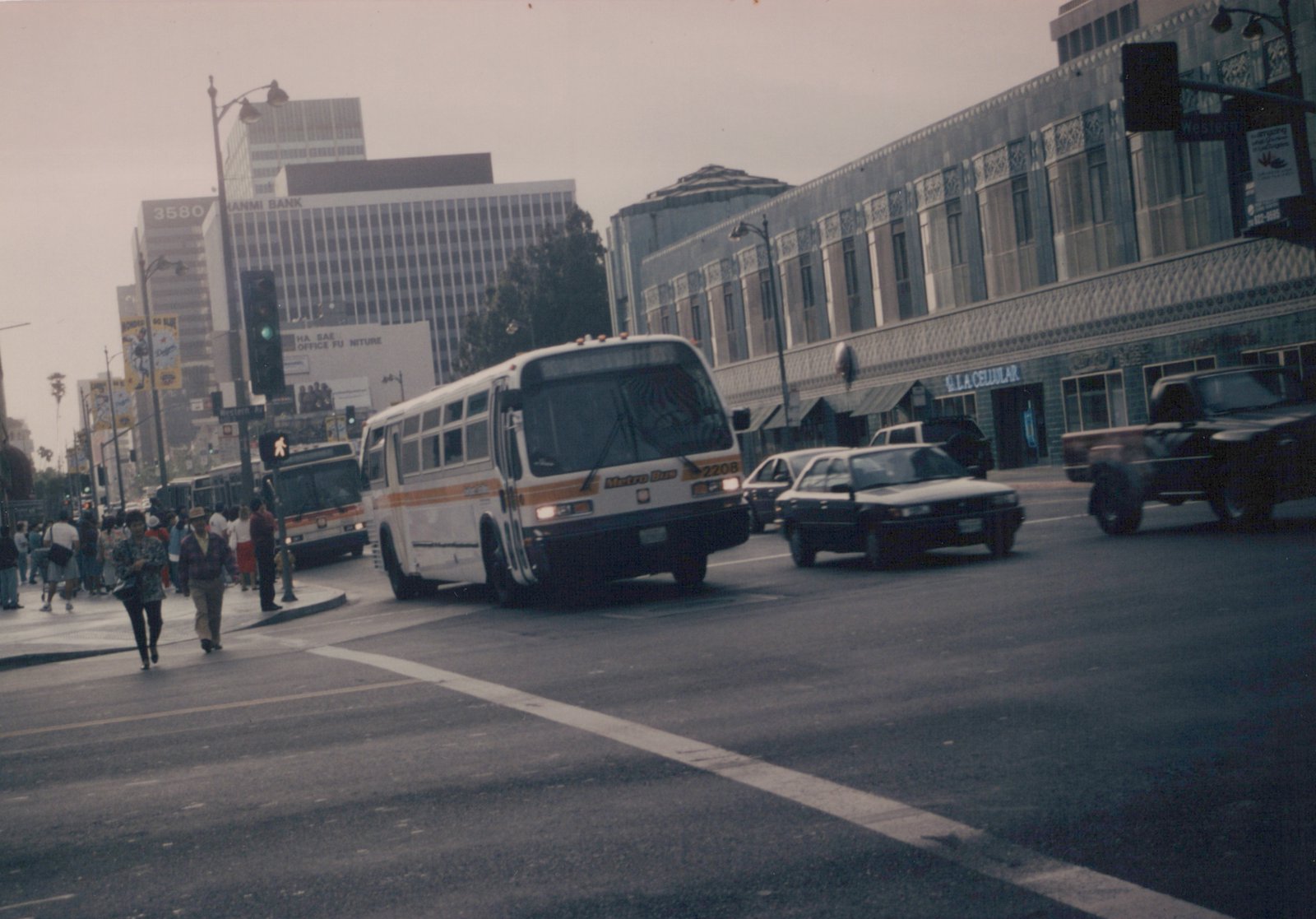A white and orange Metro Bus is turning at a city intersection with pedestrians crossing and cars nearby, under a cloudy sky.