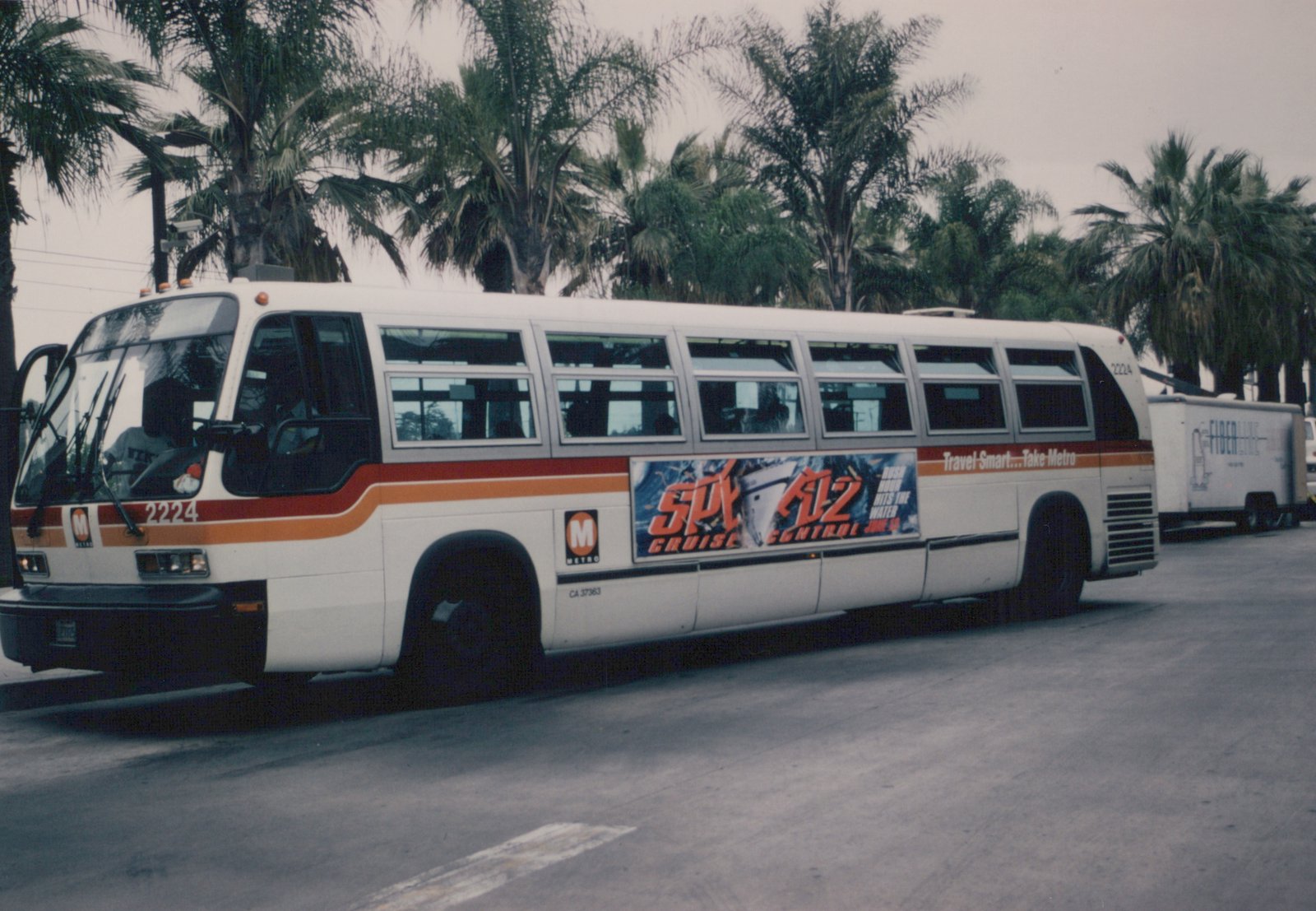 A white LA Metro bus with red and orange stripes, numbered 2224, is parked on a street lined with palm trees; it features a 'Spy Kids 2' movie ad on its side.