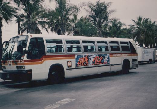 A white LA Metro bus with red and orange stripes, numbered 2224, is parked on a street lined with palm trees; it features a 'Spy Kids 2' movie ad on its side.