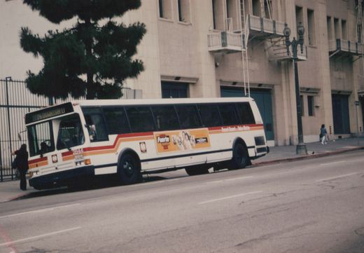 A white CTA bus with red and orange stripes, numbered 2844 and on route 26 Downtown, is parked on a street next to a sidewalk with two people and a large building in the background.
