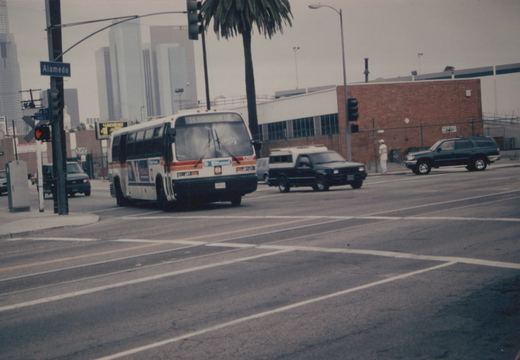 A white and orange New Look bus with the number 1237 and destination '16 DOWNTOWN LA' is stopped at a street intersection with palm trees and city buildings in the background.