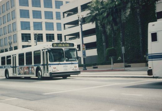 A white and blue Long Beach Transit bus, number 9720, is shown from a front-three-quarter angle stopped at an intersection on a sunny day, with modern buildings and palm trees in the background.