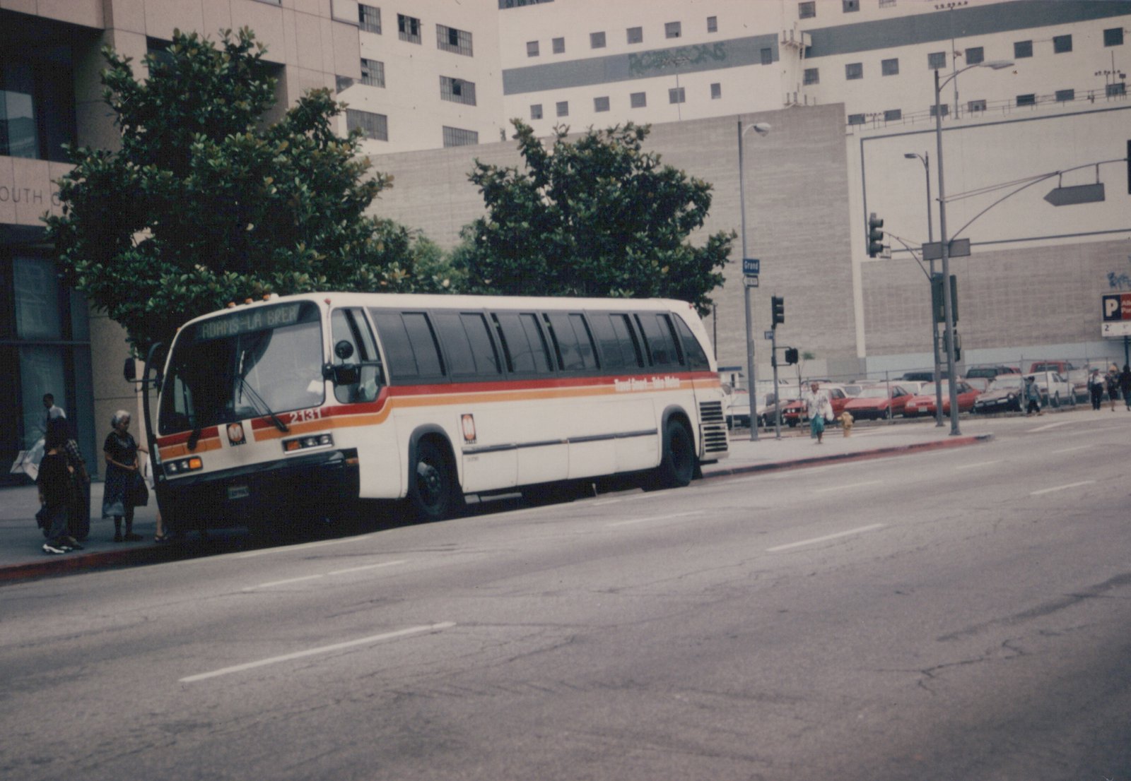 A white Flxible Metro bus with red and orange stripes, numbered 2151, is stopped on a city street with passengers nearby and a street sign reading Grand in the background.