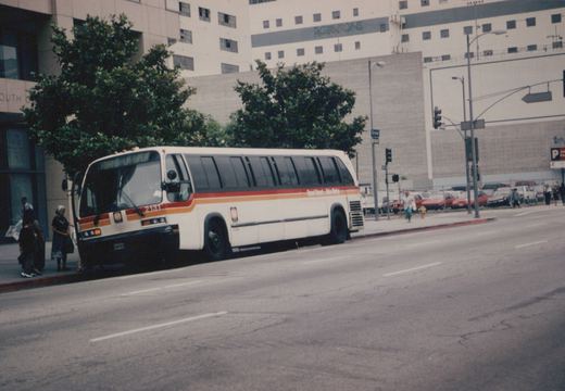 A white Flxible Metro bus with red and orange stripes, numbered 2151, is stopped on a city street with passengers nearby and a street sign reading Grand in the background.
