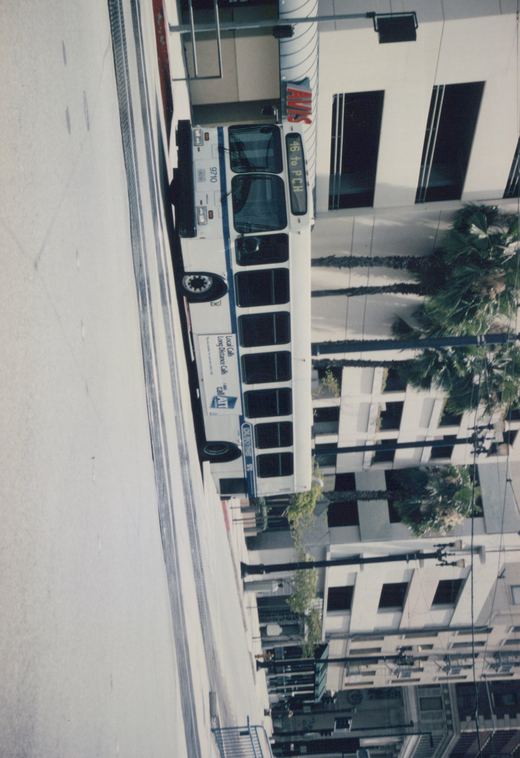 A white and blue city bus with the number 9710 and destination sign '46 to PCH' is parked on a street near a building with an 'AVIS' sign and palm trees in the background.