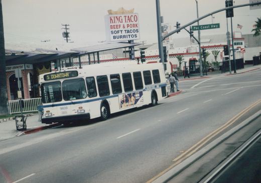 A white and blue motor bus numbered 9608 is driving on a street with a '5 DOWNTOWN' destination sign, passing a 'King Taco' restaurant and a 'Pac Coast Hwy' street sign, with a few pedestrians on the corner.
