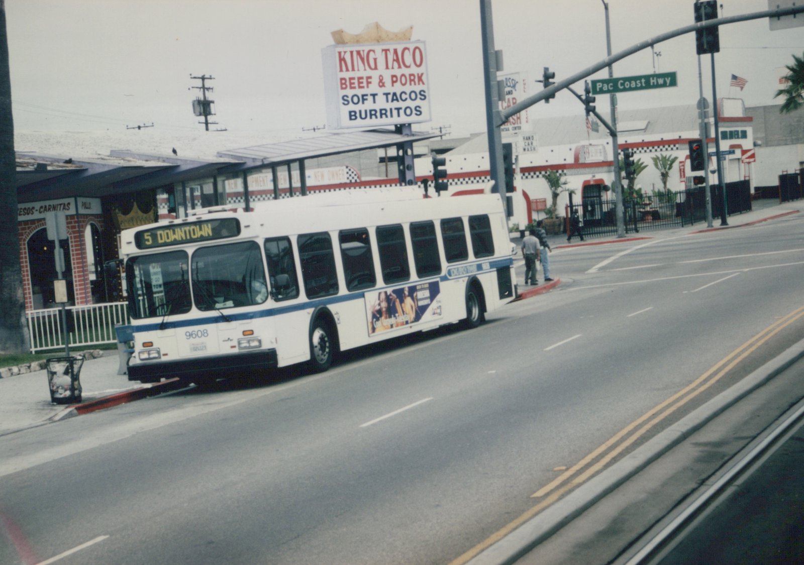 A white and blue motor bus numbered 9608 is driving on a street with a '5 DOWNTOWN' destination sign, passing a 'King Taco' restaurant and a 'Pac Coast Hwy' street sign, with a few pedestrians on the corner.