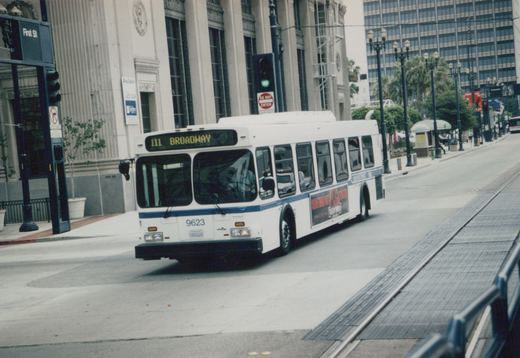 A white and blue 60-foot Metro bus, number 9623, is turning a corner on a city street, with a 'Jurassic Park' ad on its side and a 'First St' street sign visible.