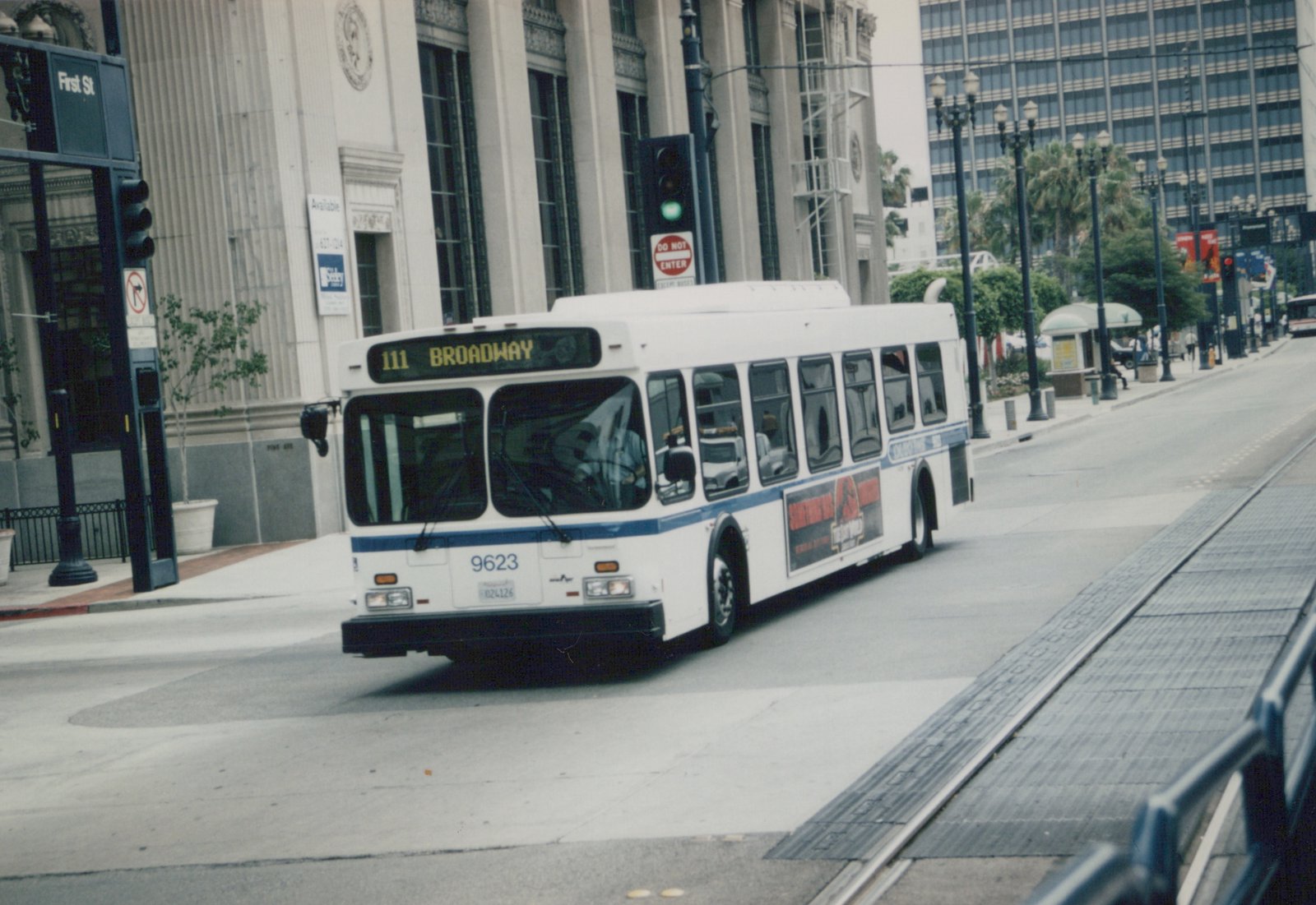 A white and blue 60-foot Metro bus, number 9623, is turning a corner on a city street, with a 'Jurassic Park' ad on its side and a 'First St' street sign visible.