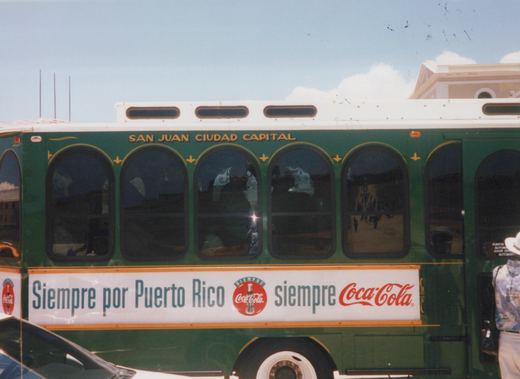 A green and white bus with 'San Juan Ciudad Capital' lettering and a 'Siempre por Puerto Rico siempre Coca-Cola' ad on its side, parked on a street with people inside and a person standing beside it.