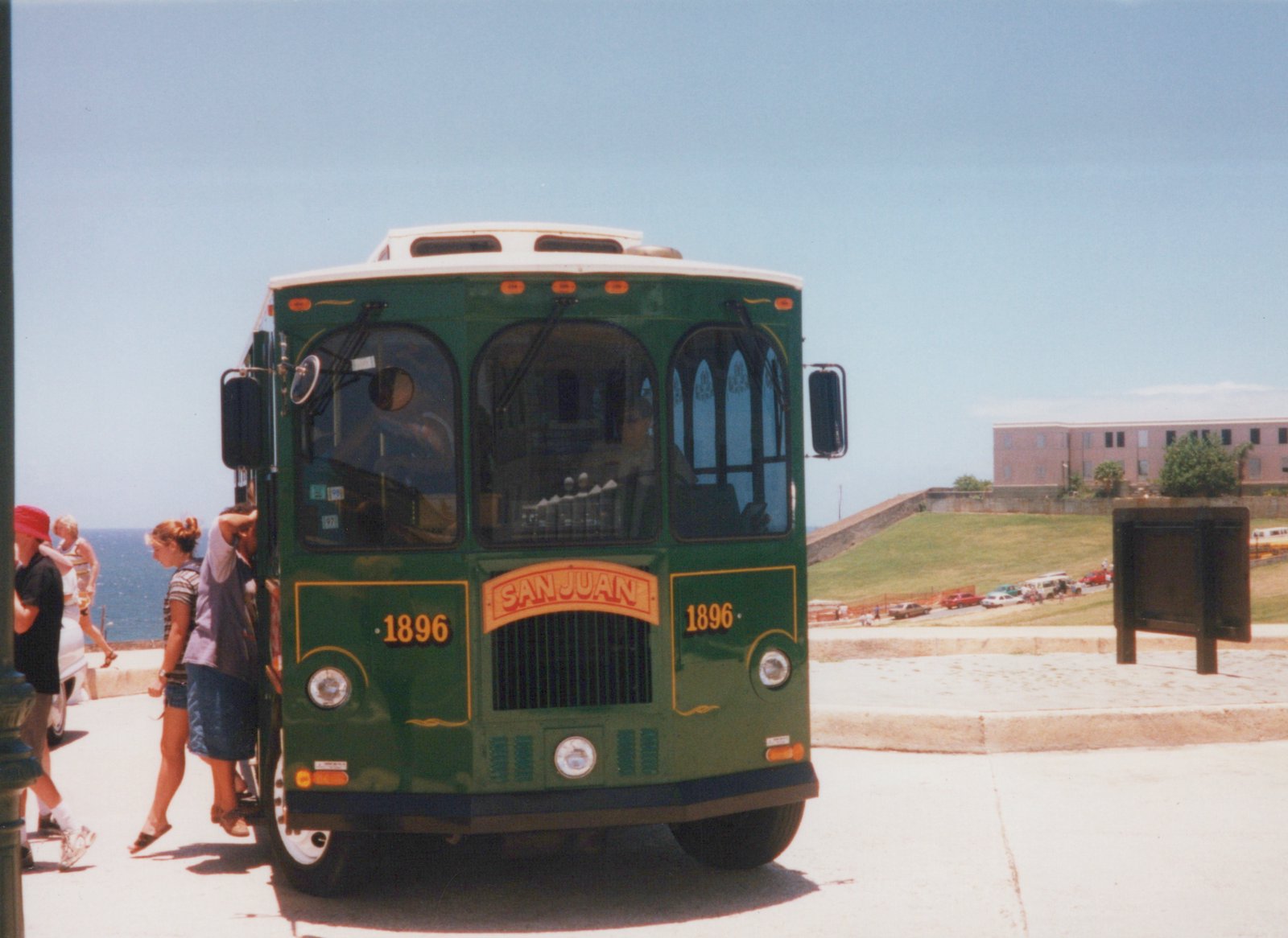 Front view of a green San Juan trolleybus, number 1896, with passengers boarding in a sunny, coastal setting.