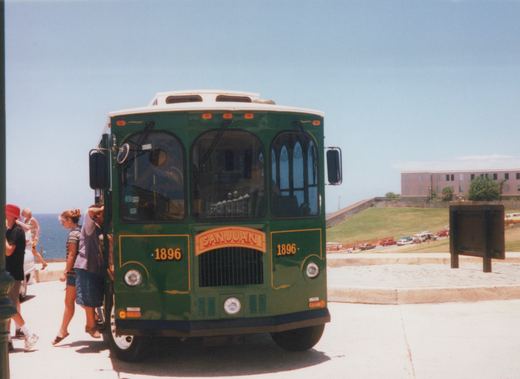 Front view of a green San Juan trolleybus, number 1896, with passengers boarding in a sunny, coastal setting.