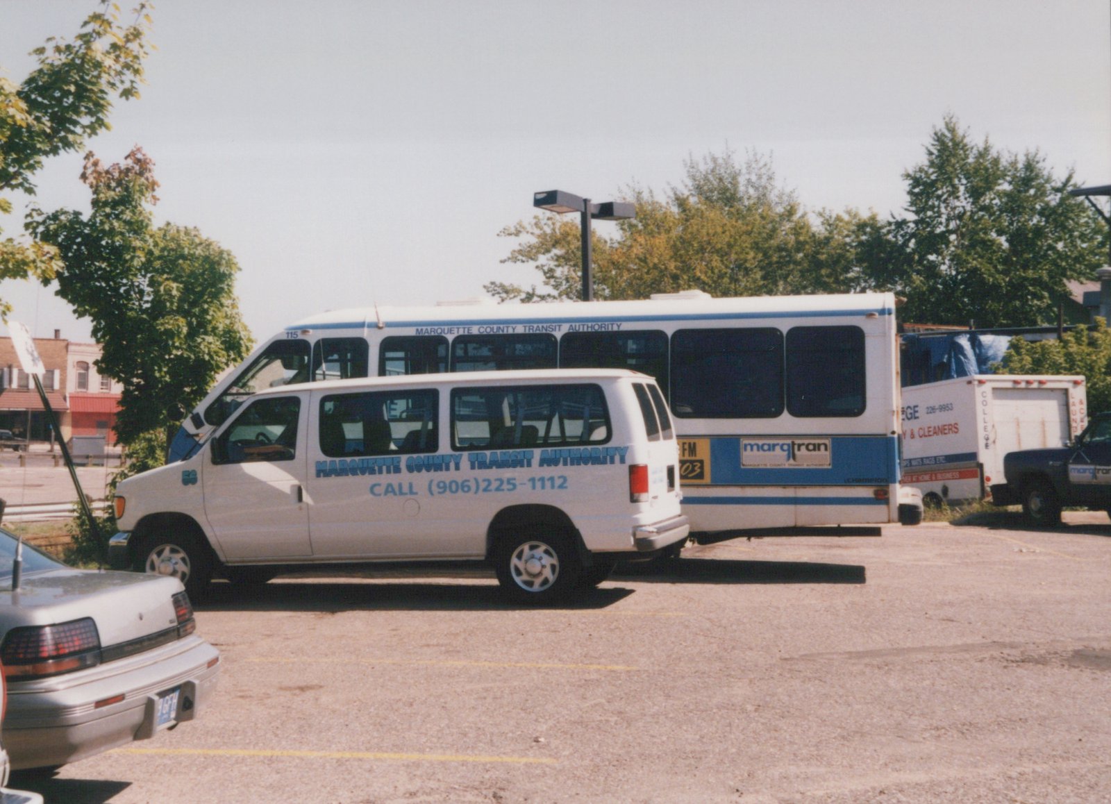 A white and blue Marquette County Transit Authority bus and a matching white van are parked together in a paved lot on a sunny day in Marquette, Michigan.