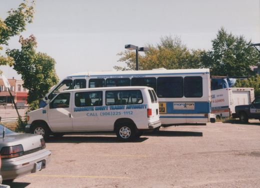 A white and blue Marquette County Transit Authority bus and a matching white van are parked together in a paved lot on a sunny day in Marquette, Michigan.