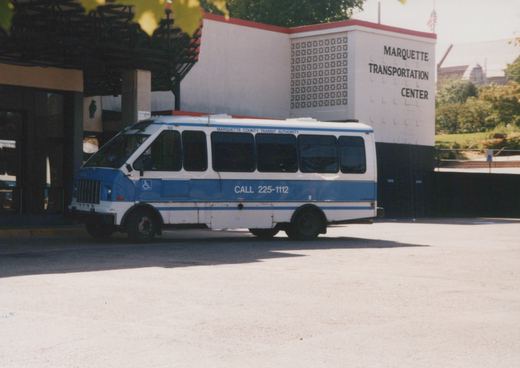 A side view of a white and blue Marquette County Transit Authority GMC bus number 50 parked in front of the Marquette Transportation Center building under a clear sky.