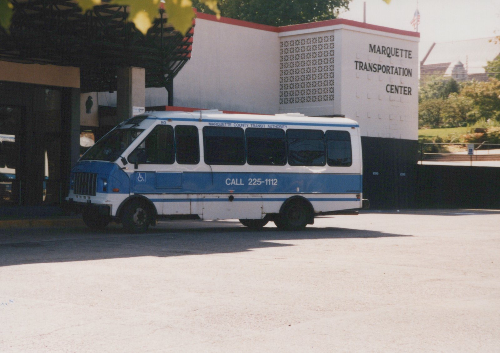A side view of a white and blue Marquette County Transit Authority GMC bus number 50 parked in front of the Marquette Transportation Center building under a clear sky.