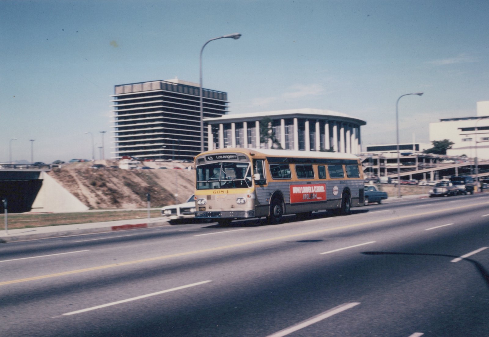 A yellow and silver bus with the number 6054 and destination 'Los Angeles' drives on a multi-lane road under a clear blue sky, with large buildings in the background.