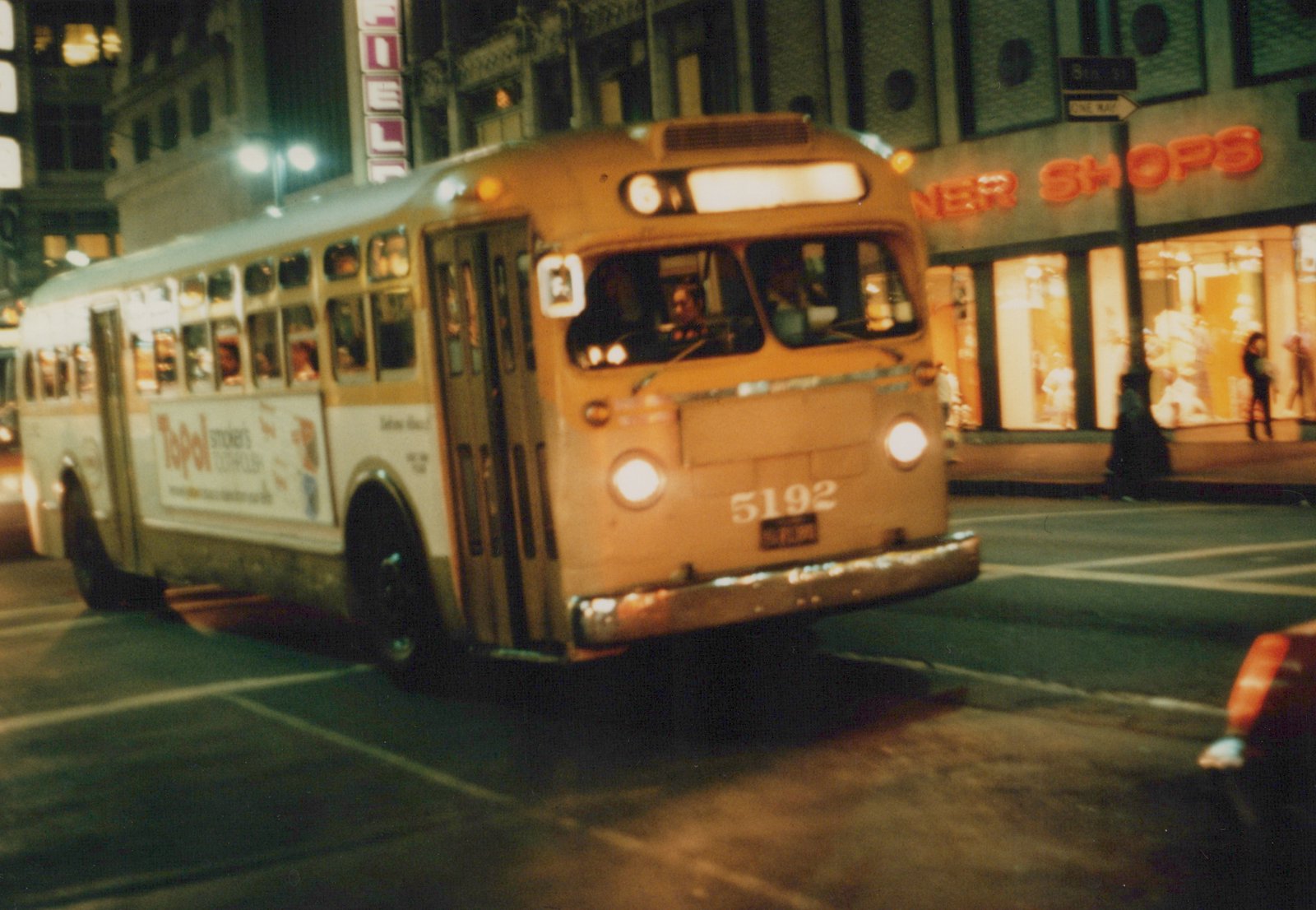 A yellow and white CTA bus with the number 5192 on the front, captured at night on a city street with illuminated storefronts and a street sign for Broadway in the background.