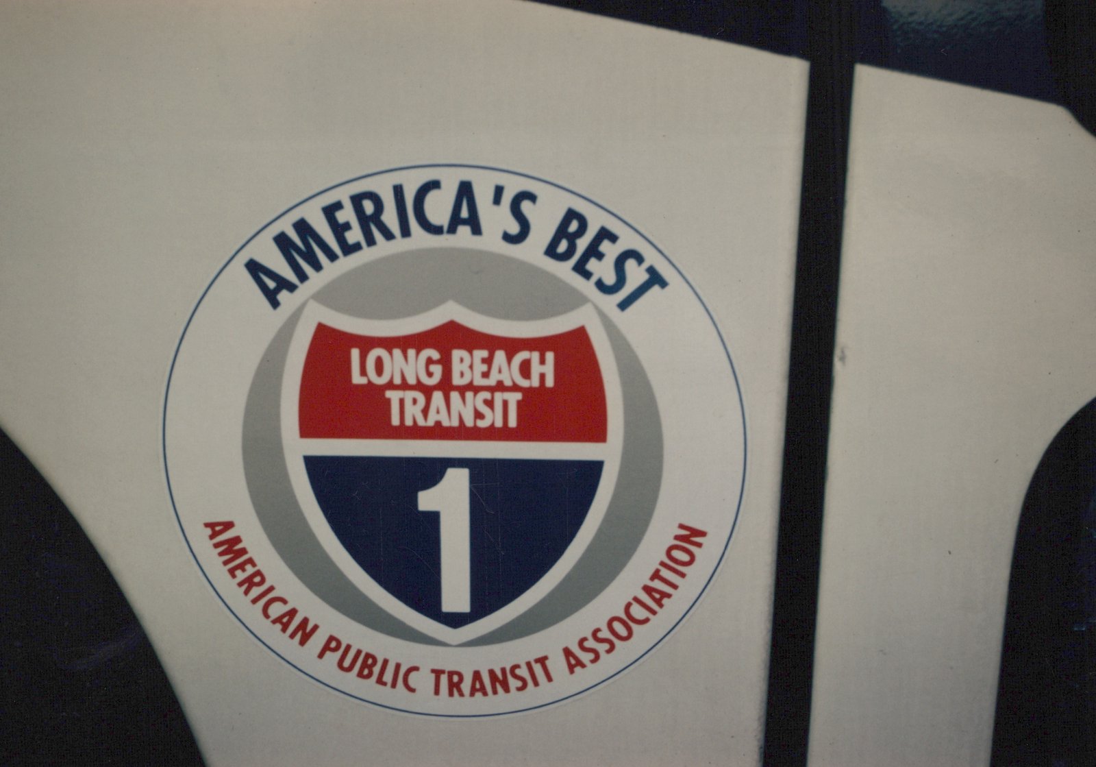 A circular decal on a white surface reading 'AMERICA'S BEST' at the top, 'LONG BEACH TRANSIT' over a red shield, a large '1' on a blue background, and 'AMERICAN PUBLIC TRANSIT ASSOCIATION' at the bottom.