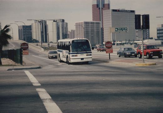 A white and blue motor bus is driving on a city street with modern buildings in the background and several cars around it.
