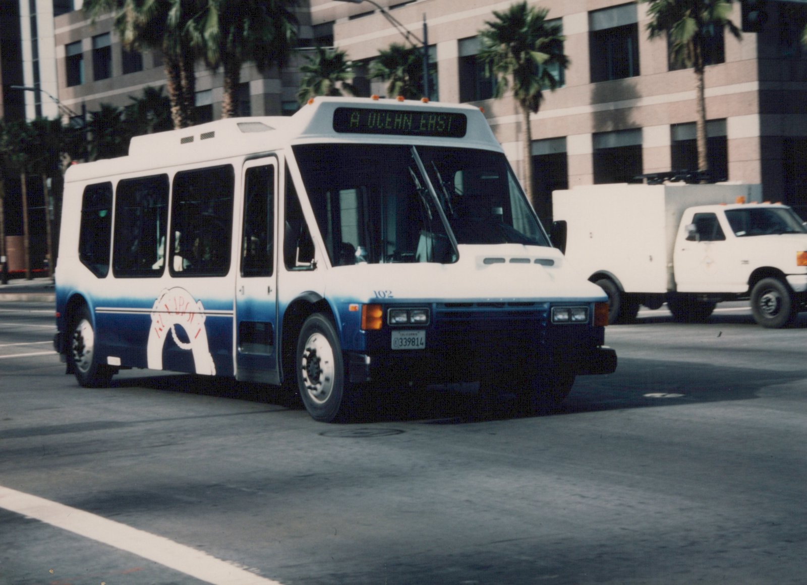 A white and blue Metro bus with the number 102 and destination 'A OCEAN EAST' is driving down a city street lined with palm trees and a modern building in the background.