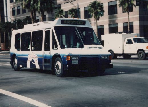 A white and blue Metro bus with the number 102 and destination 'A OCEAN EAST' is driving down a city street lined with palm trees and a modern building in the background.
