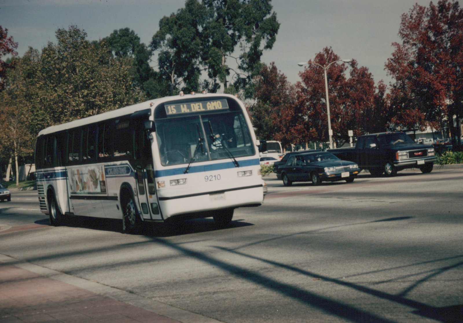 A white and blue bus with the number 9210 and destination sign '15 W. DEL AMO' is driving down a city street lined with trees showing autumn colors.