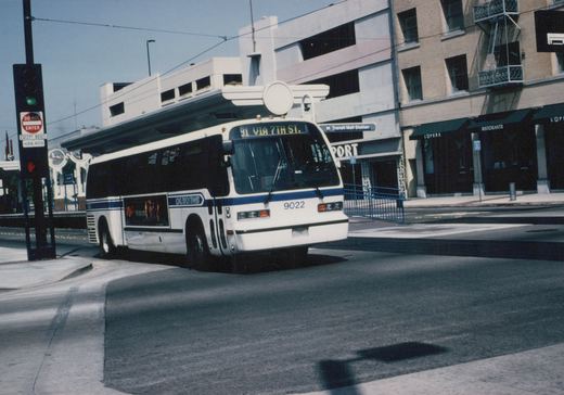 A white and blue Long Beach Transit bus numbered 9022 is turning on a city street with a sign for M Transit Mall Station and storefronts in the background.