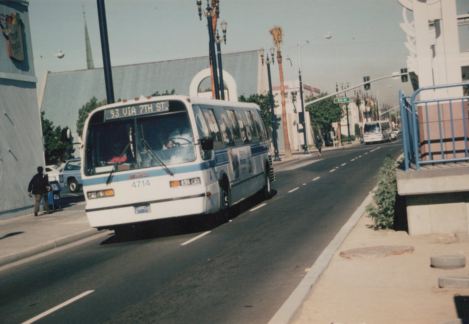 White and blue bus with route 93 via 7th St on its sign, driving down a street with palm trees and a '5th St' sign visible, with another similar bus in the background.