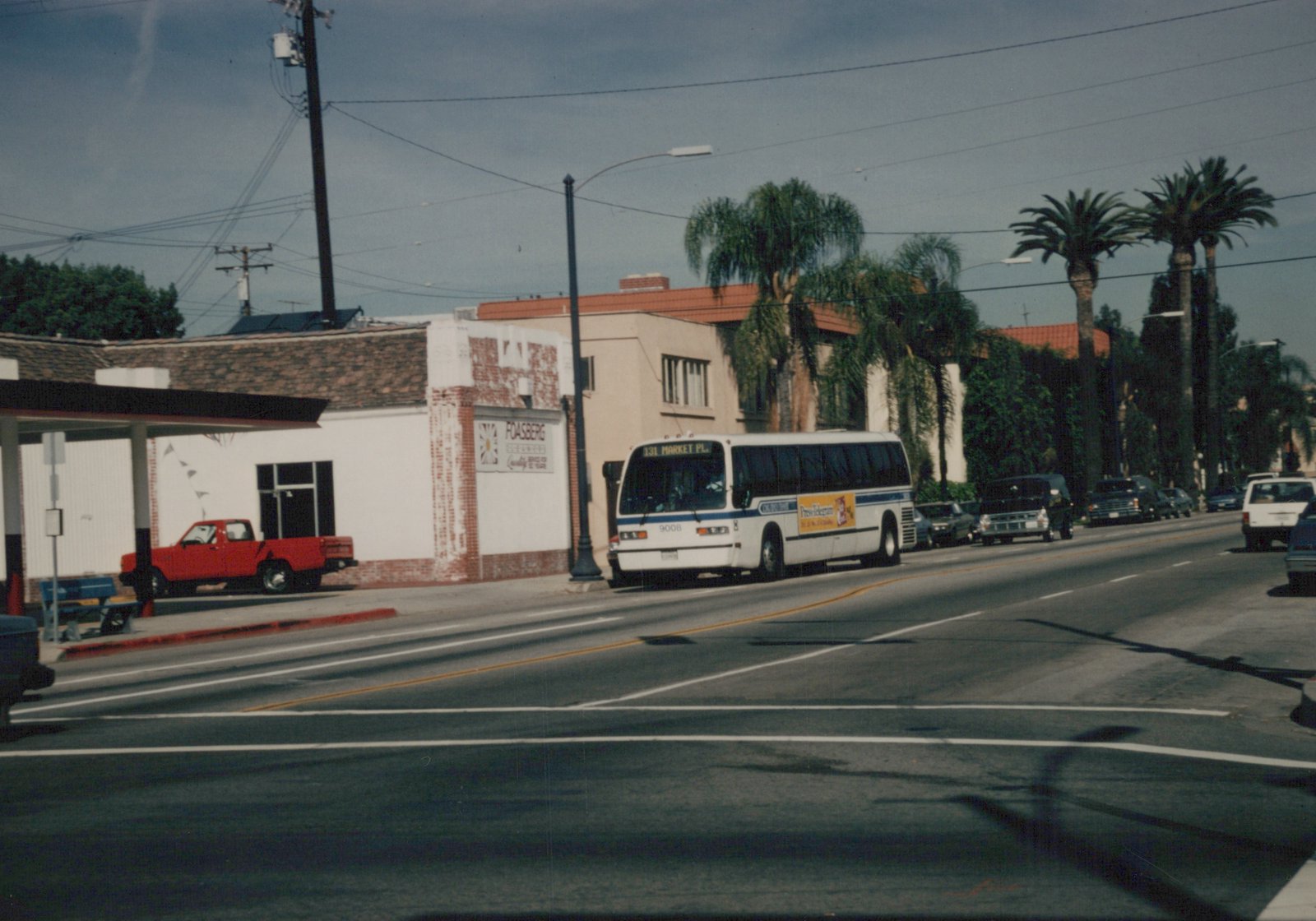 A white and blue motor bus with the number 9008 and a destination sign reading '131 Market Pl.' is stopped on a sunny street lined with palm trees and parked cars.