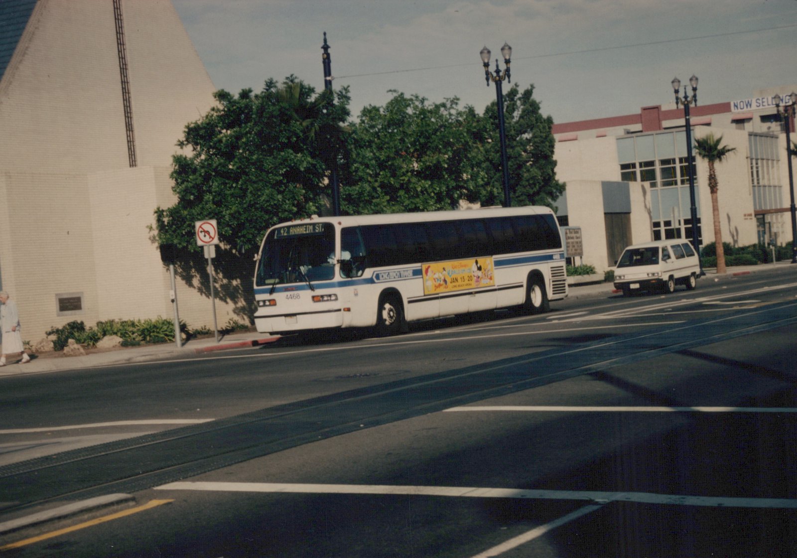 A white Long Beach Transit bus with blue stripes, marked 4468 and route 42 to Anaheim St, drives down a city street past a van and a building with a 'NOW SELLING' sign.