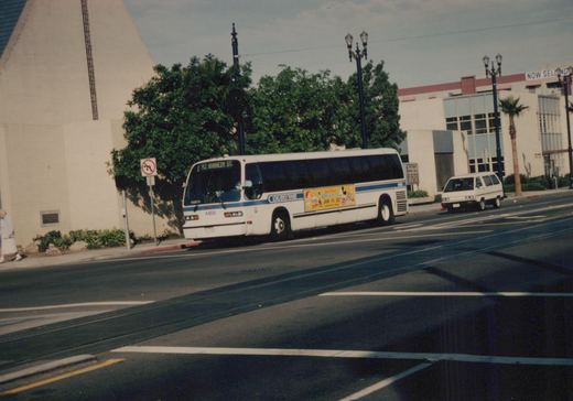 A white Long Beach Transit bus with blue stripes, marked 4468 and route 42 to Anaheim St, drives down a city street past a van and a building with a 'NOW SELLING' sign.
