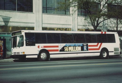 Side view of a white city bus with red stripes and a Kentucky Lottery ad, parked on a city street with a modern building in the background.