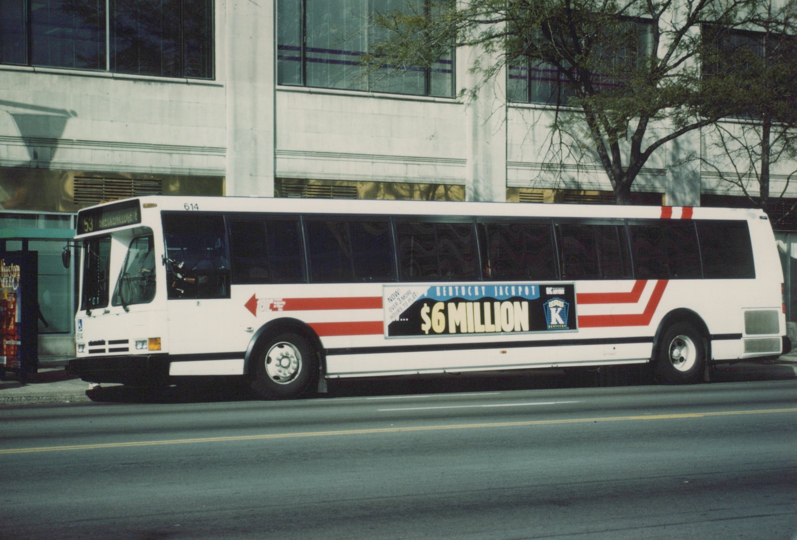 Side view of a white city bus with red stripes and a Kentucky Lottery ad, parked on a city street with a modern building in the background.
