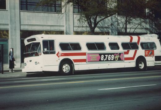 A white Flxible Metro bus with red stripes and a lottery ad on its side is parked on a city street, with a person waiting at a nearby bus stop.