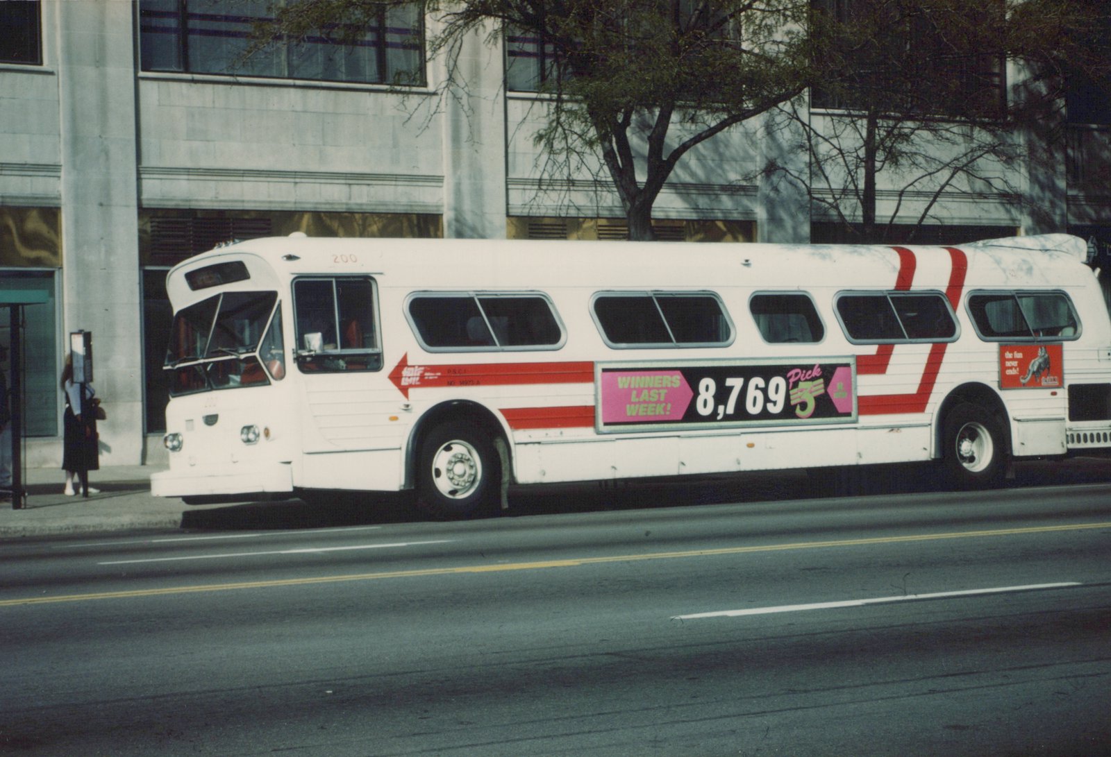 A white Flxible Metro bus with red stripes and a lottery ad on its side is parked on a city street, with a person waiting at a nearby bus stop.