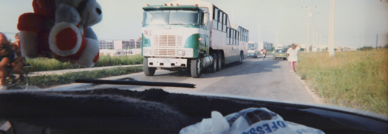 A view from inside a car shows a green and white MI bus, number 3570, driving on a road with tall grass on both sides, with a person standing on the right and another on a bicycle on the left.