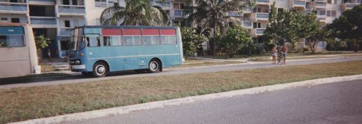 Side view of a vintage light blue Flxible bus with a red band, parked on a paved road next to a grassy area, with a multi-story apartment building and palm trees in the background.