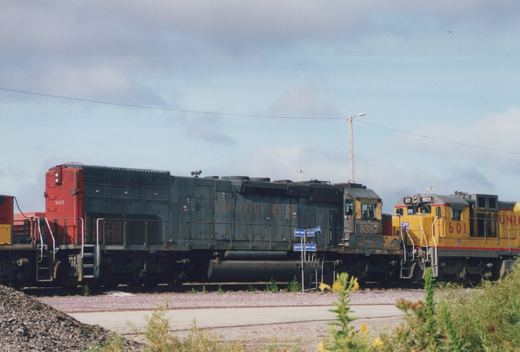 Two diesel locomotives, one blue and red with the number 9387 and the words 'COTTON BELT' on its side, and the other yellow with the number 601 and partial 'UNION PACIFIC' lettering, are parked on parallel tracks in a gravel yard with some greenery in the foreground.