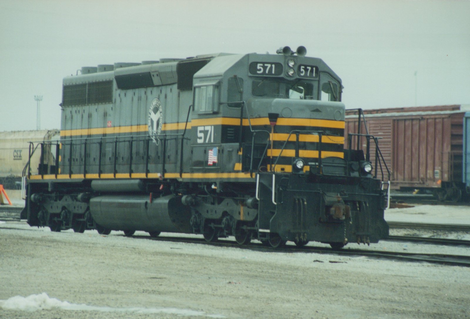 A dark gray and yellow diesel locomotive with the number 571 and a 'Y' logo, standing on tracks in a yard with snow on the ground.