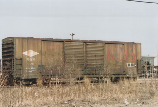 A rusty brown freight car marked MNS 909 is parked in a weed-choked yard under a grey sky.
