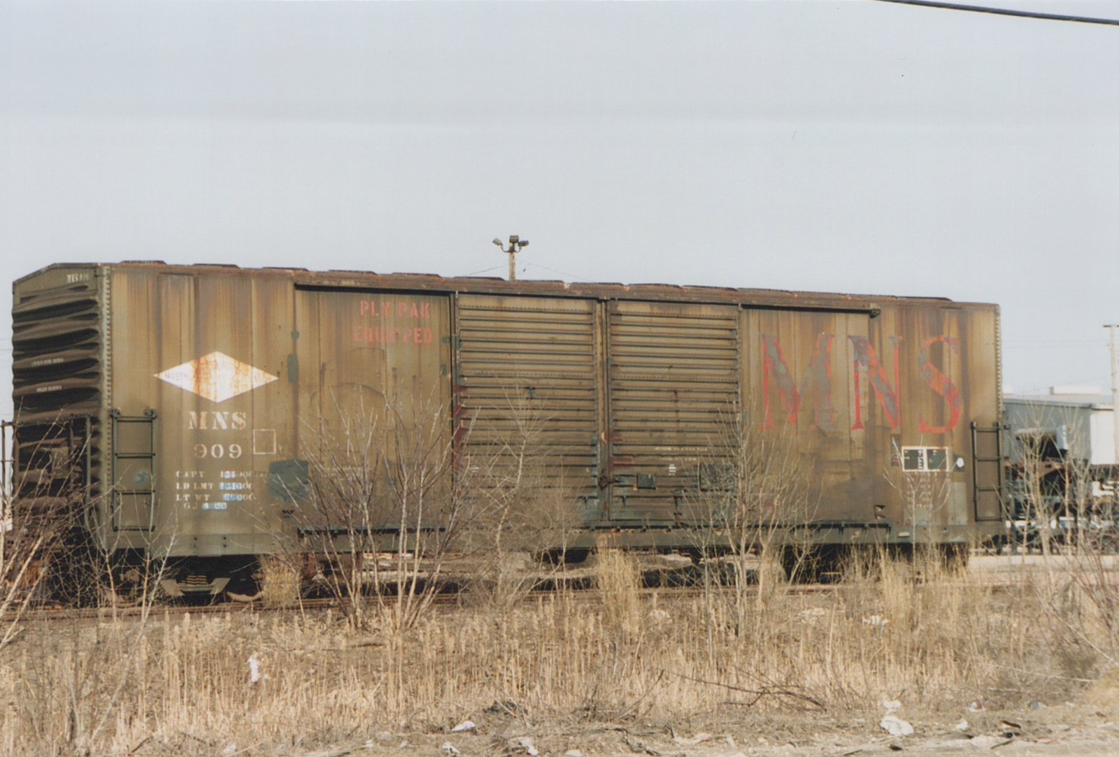 A rusty brown freight car marked MNS 909 is parked in a weed-choked yard under a grey sky.