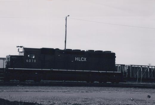 A black HLCX diesel locomotive numbered 6078 is parked on tracks in a gravel yard under an overcast sky.