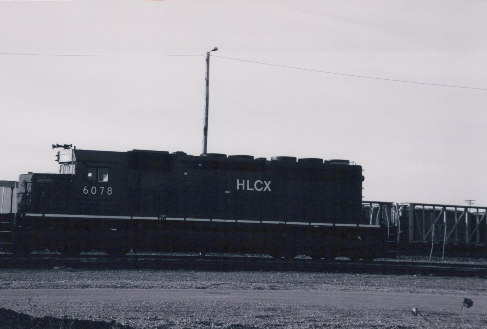 A black HLCX diesel locomotive numbered 6078 is parked on tracks in a gravel yard under an overcast sky.