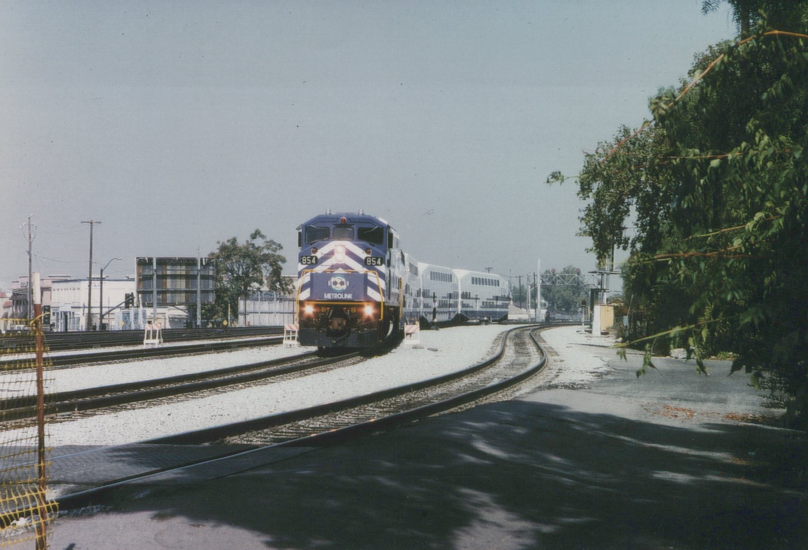 A blue and white Metrolink train with locomotive 854 travels on tracks beside a road, with buildings and trees in the background.