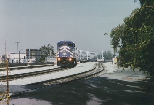A blue and white Metrolink train with locomotive 854 travels on tracks beside a road, with buildings and trees in the background.