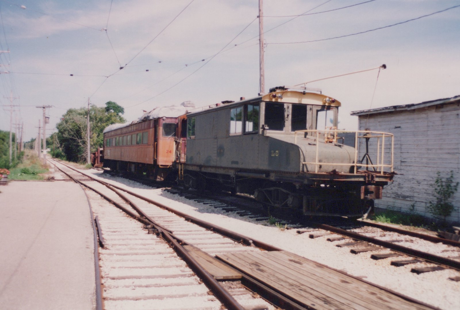 A gray electric locomotive with 'L S' lettering and an orange passenger car numbered 24 are coupled and parked on parallel tracks in a sunny yard with overhead wires and a white building nearby.