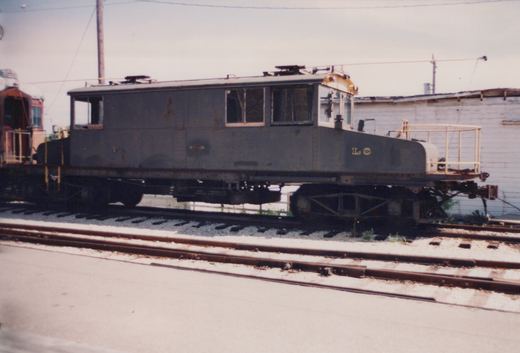 A grey, weathered electric railcar with 'LS' on its side and trolley poles on its roof is parked on railroad tracks next to a white building, with a red car partially visible behind it.
