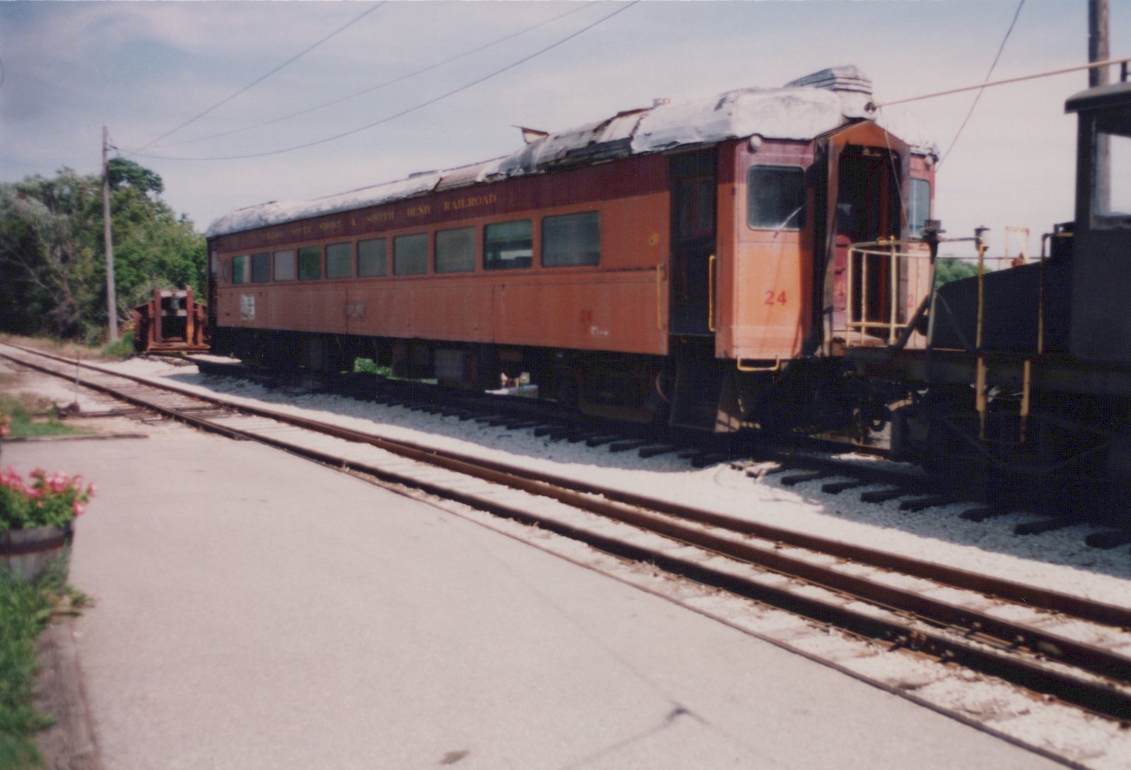 A vintage orange and maroon interurban railcar, numbered 24 and lettered for the Chicago South Shore and South Bend Railroad, is parked on tracks in a sunny yard with a damaged roof.